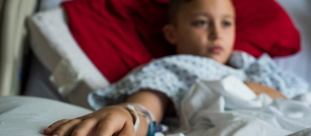"Child lying in hospital bed with IV in hand, wearing hospital gown"